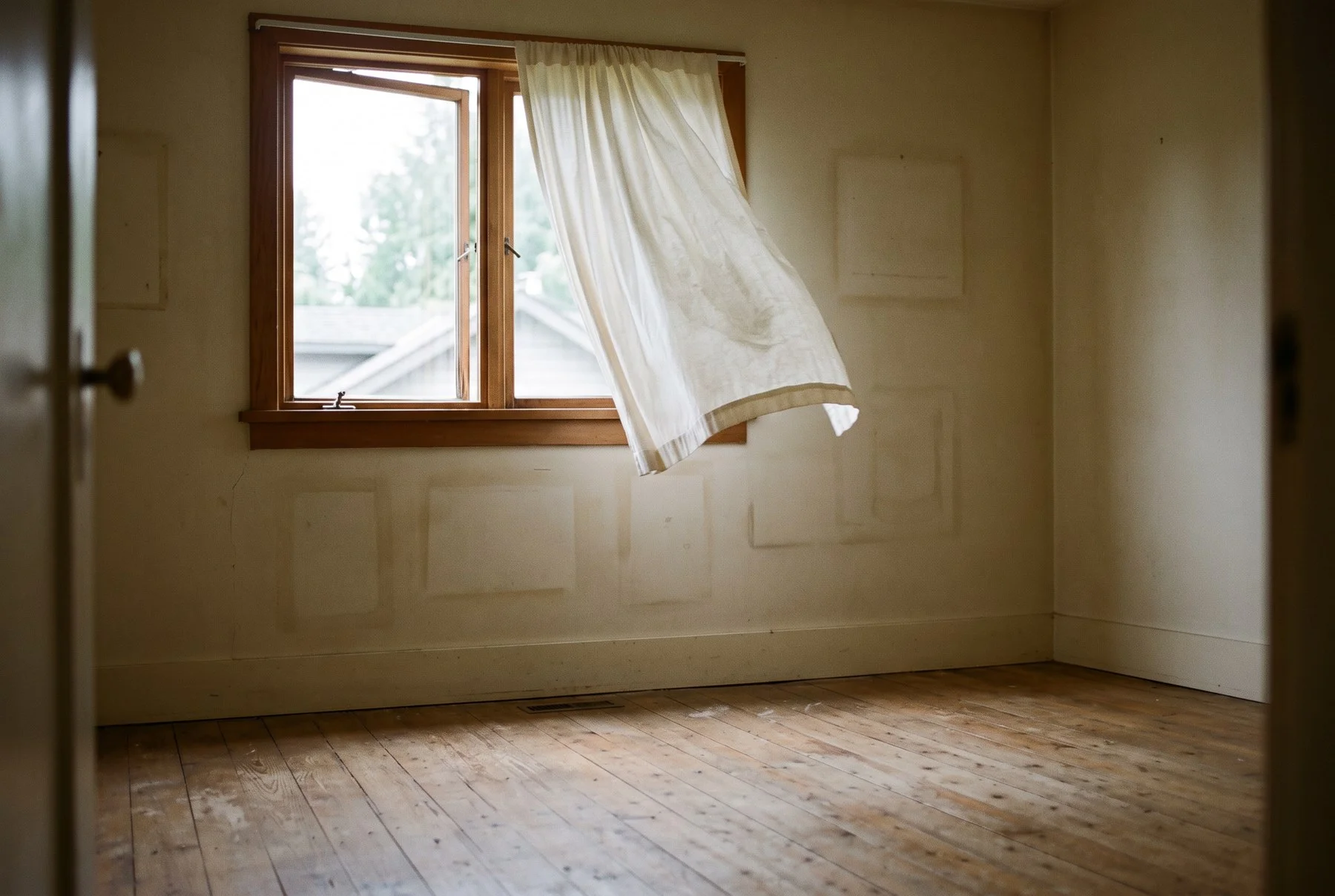 An empty rental bedroom with bare wood floors and a curtain blowing at an open window after move-out