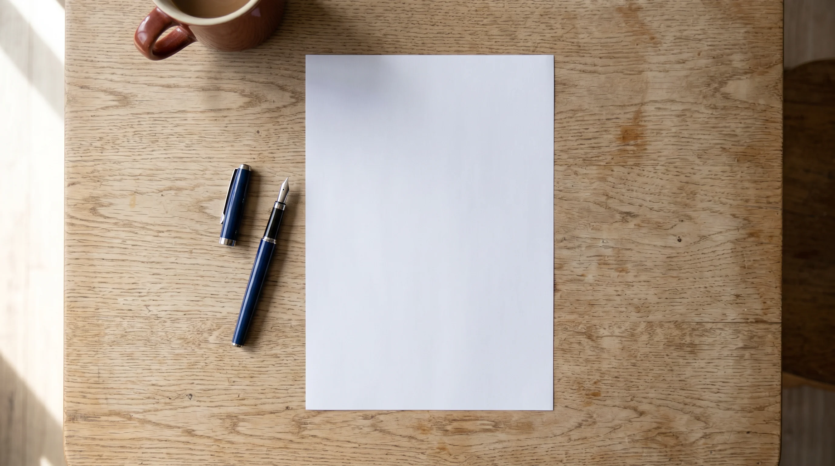 A blank sheet of white paper on a pale wood kitchen table with an uncapped fountain pen beside it, in soft morning daylight. Nothing is printed on the page.